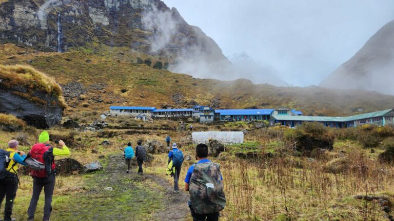 Trekkers with backpacks head toward blue-roofed lodges, ideal for trekking in Nepal's Annapurna Base Camp trek 2025