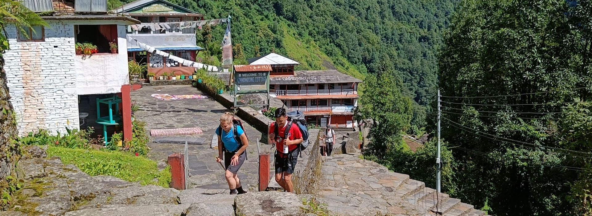 High-altitude village entry with trekkers climbing under sunny skies