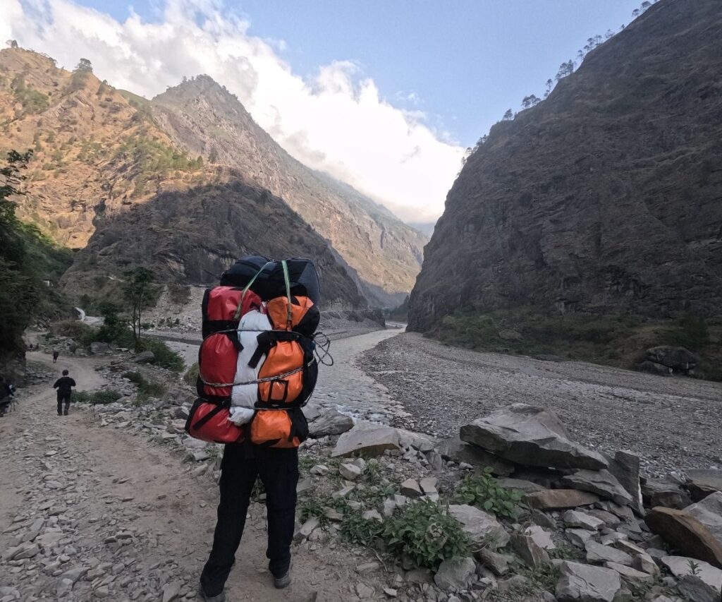 trek porter walking beside a river with large backpacks tied together