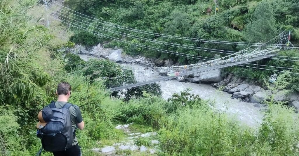 Trekker approaching a suspension bridge over a fast-flowing river in the lush green hills of Nepal
